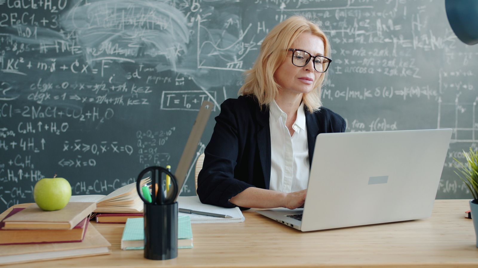 Lehrkraft mit Brille arbeitet im Klassenzimmer an einem Laptop, im Hintergrund eine Tafel mit Formeln